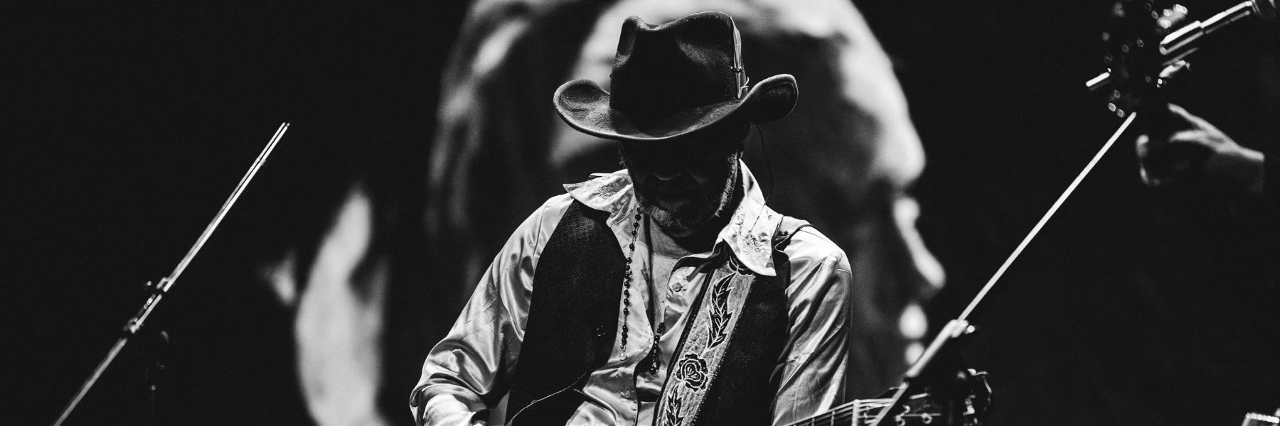 Guitarist in a cowboy hat and vest playing an electric guitar on stage in black and white.