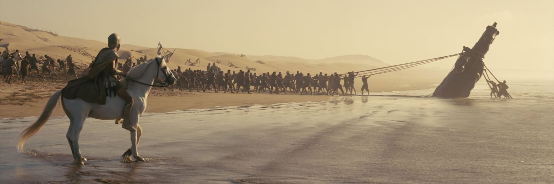 A long line of people pulling a large ship ashore by ropes as a rider on a white horse watches from the foreground.