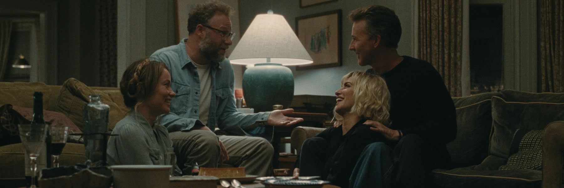 Four friends chat and laugh in a cozy living room, sitting on a sofa and the floor around a coffee table.