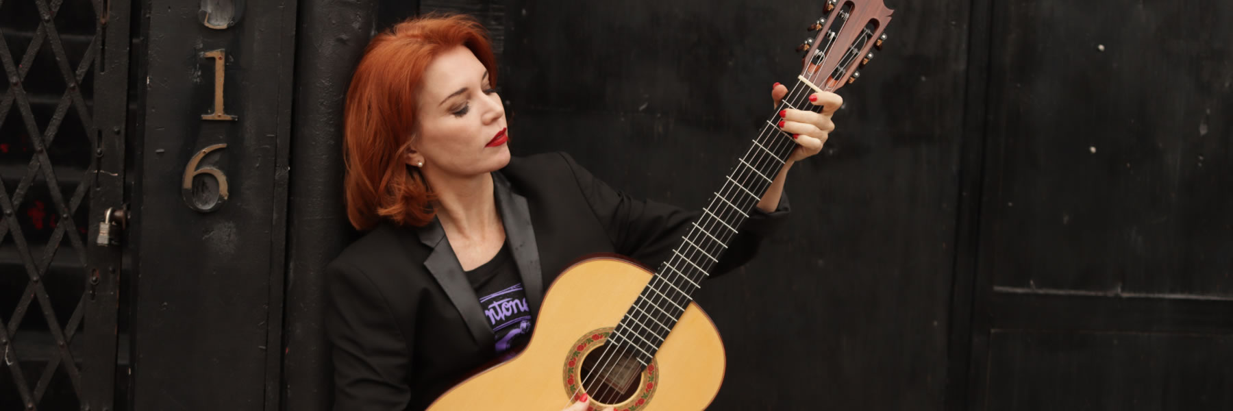 Woman with red hair in a black blazer playing a classical guitar against a dark background, looking down at the strings.