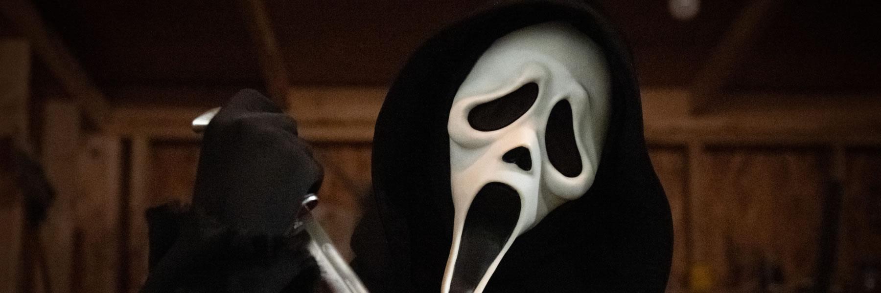 Hooded figure wearing a Ghostface mask and black cloak, holding a knife in a dimly lit room.
