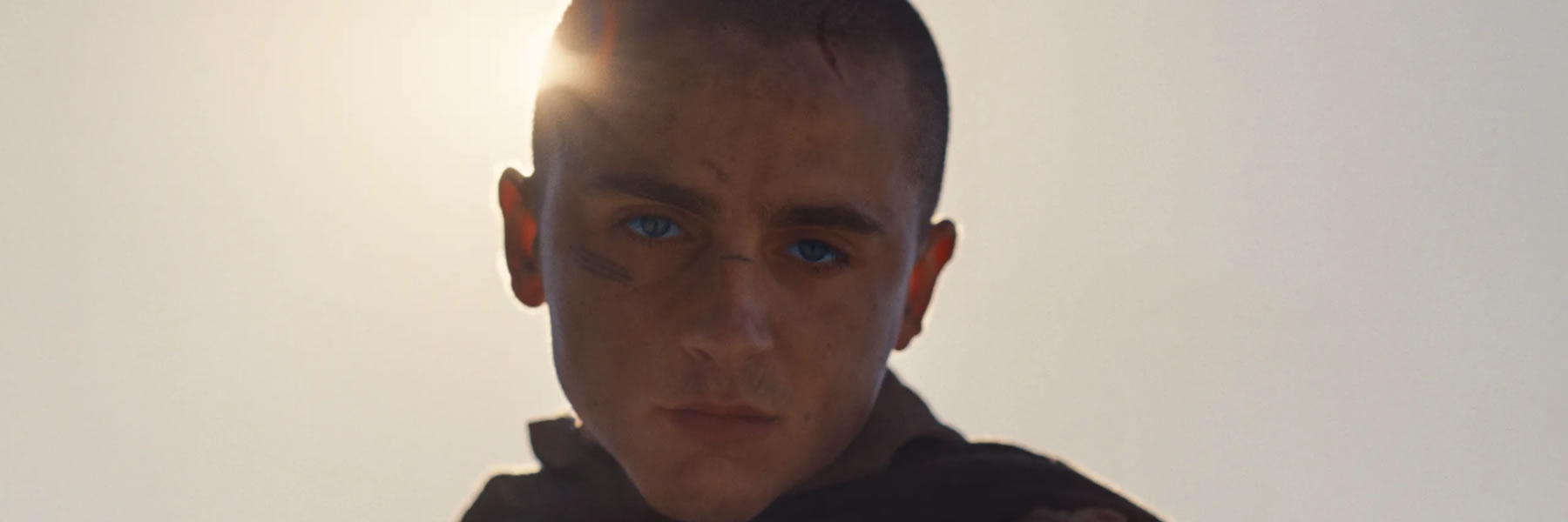 Close-up of a young man with a shaved head and blue eyes, a serious, determined expression, sun behind him.