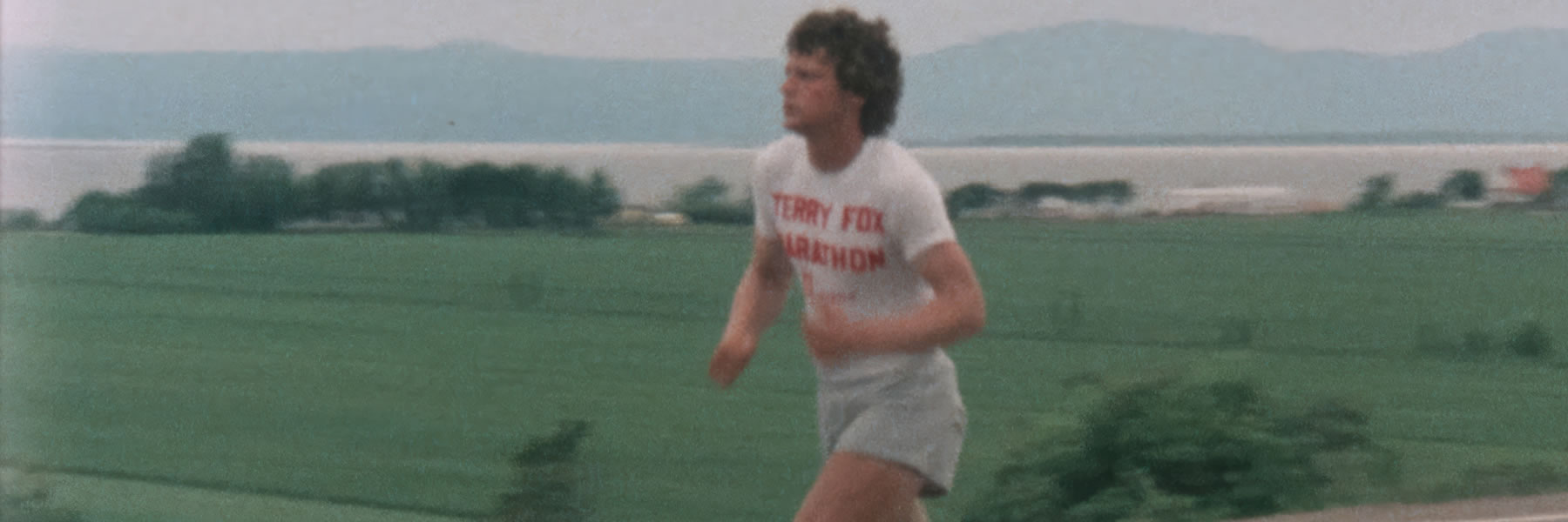 Runner in a white T-shirt reading 'TERRY FOX MARATHON' sprinting across a grassy field with hills in the background on a cloudy day.