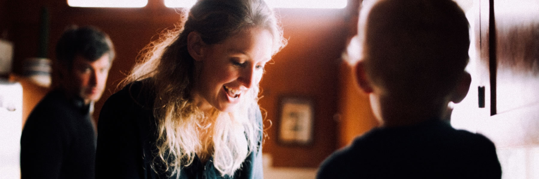 Woman with long blonde hair smiling and leaning toward someone off-camera in a warmly lit room, with a man in the background.